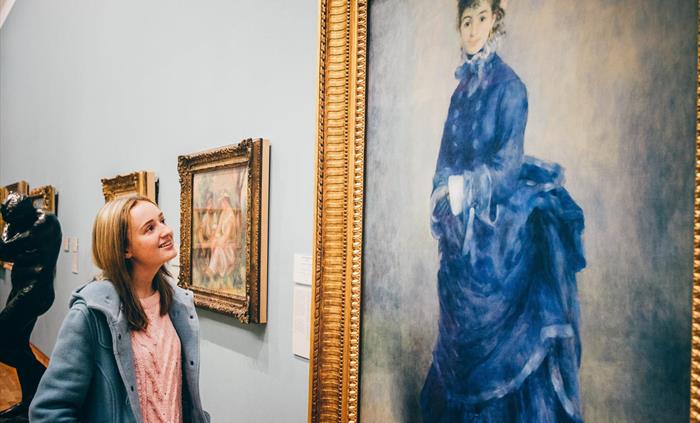 young lady looking at painting by Renoir called La Parisienne. Painting shows a young woman wearing a blue dress.